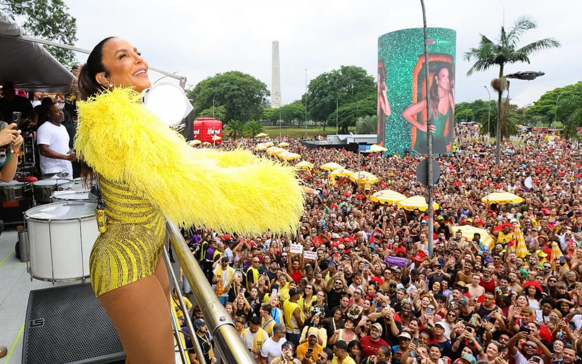 Ivete Sangalo vista de costas em cima de um trio elétrico, usando um figurino amarelo com franjas, diante de uma multidão em um bloco de Carnaval.