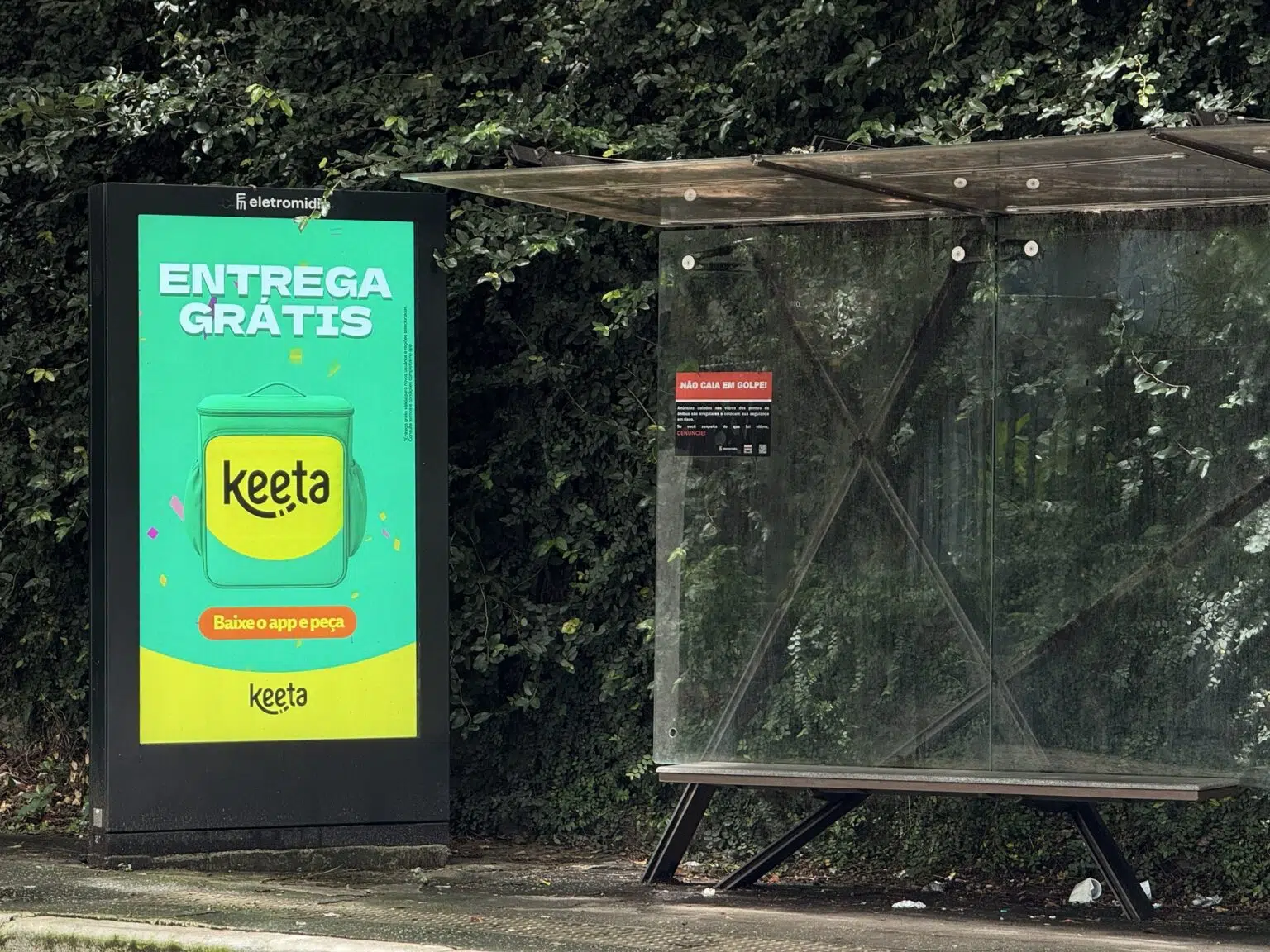 Totem publicitário digital em um ponto de ônibus exibindo um anúncio da Keeta com a frase "ENTREGA GRÁTIS" sobre um fundo verde vibrante e a imagem da mochila de entrega da marca.