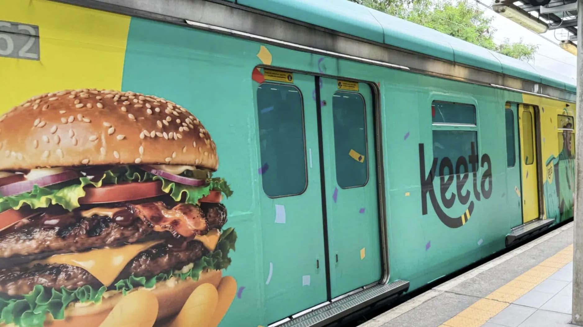 Lateral de um vagão de metrô totalmente adesivado com a identidade visual da Keeta, exibindo um hambúrguer realista gigante e o logotipo da marca em um fundo verde-água.