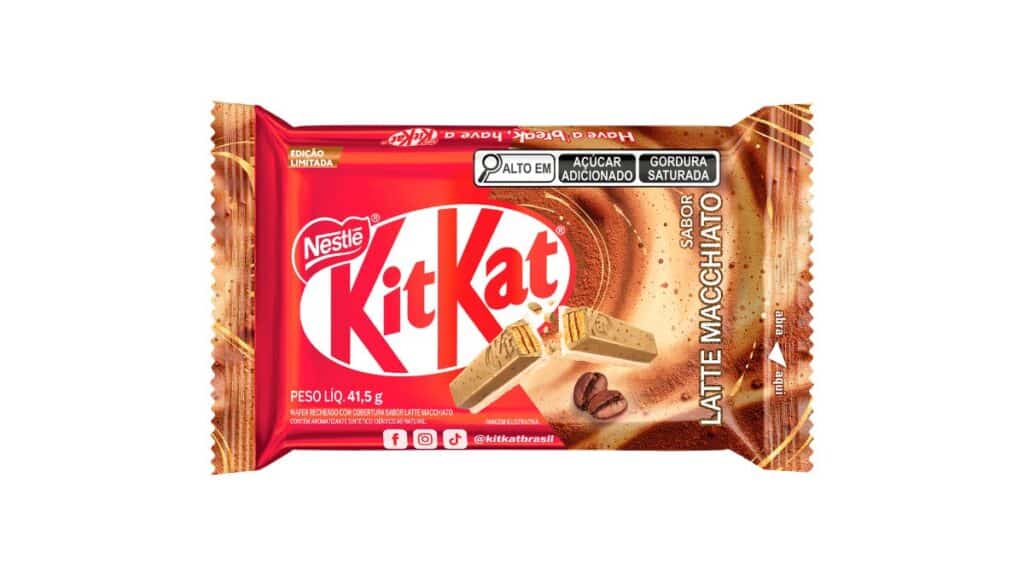 : Embalagem de KitKat sabor Latte Macchiato sobre uma mesa de madeira, ao lado de uma xícara de café fumegante e grãos espalhados.