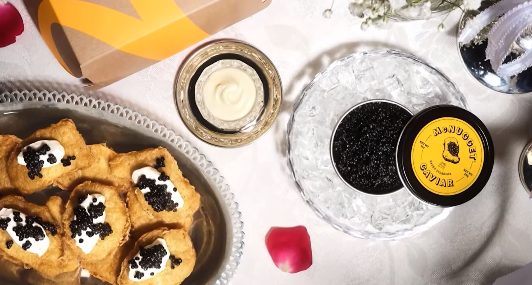 Close-up de uma bandeja de prata com McNuggets cobertos com creme branco e caviar, ao lado de uma lata aberta de "McNugget Caviar" repousando sobre gelo.