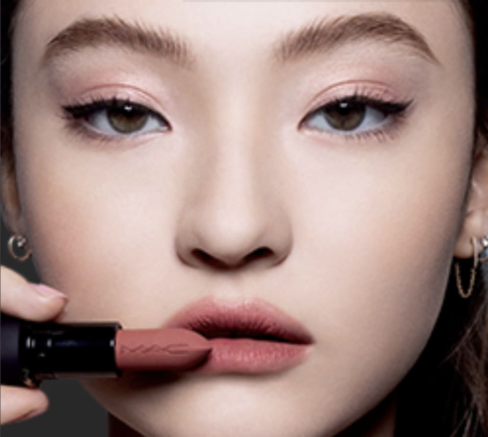 Close-up do rosto de uma modelo de pele clara aplicando um batom em bala de tom marrom rosado nos lábios.