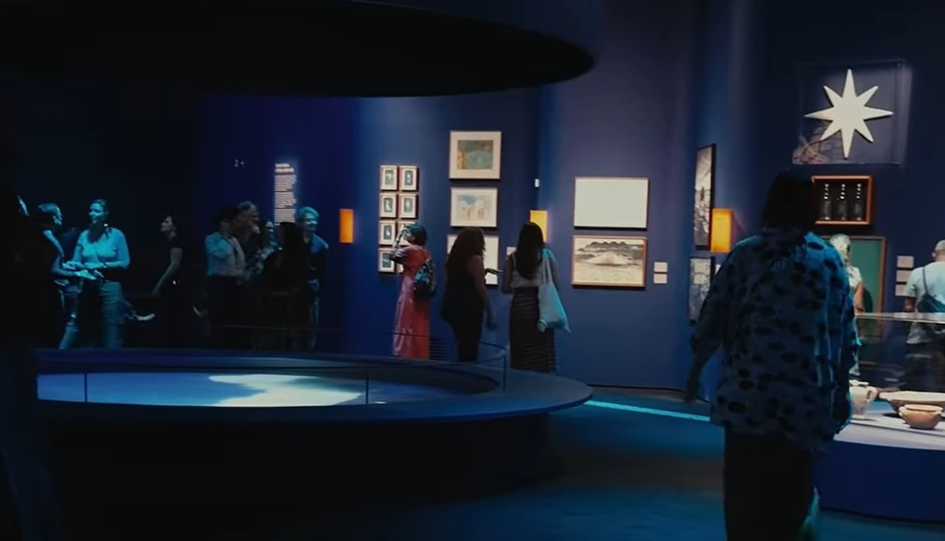 Visitantes em uma galeria de museu com paredes azul-escuro, observando quadros e objetos expostos sob iluminação suave.