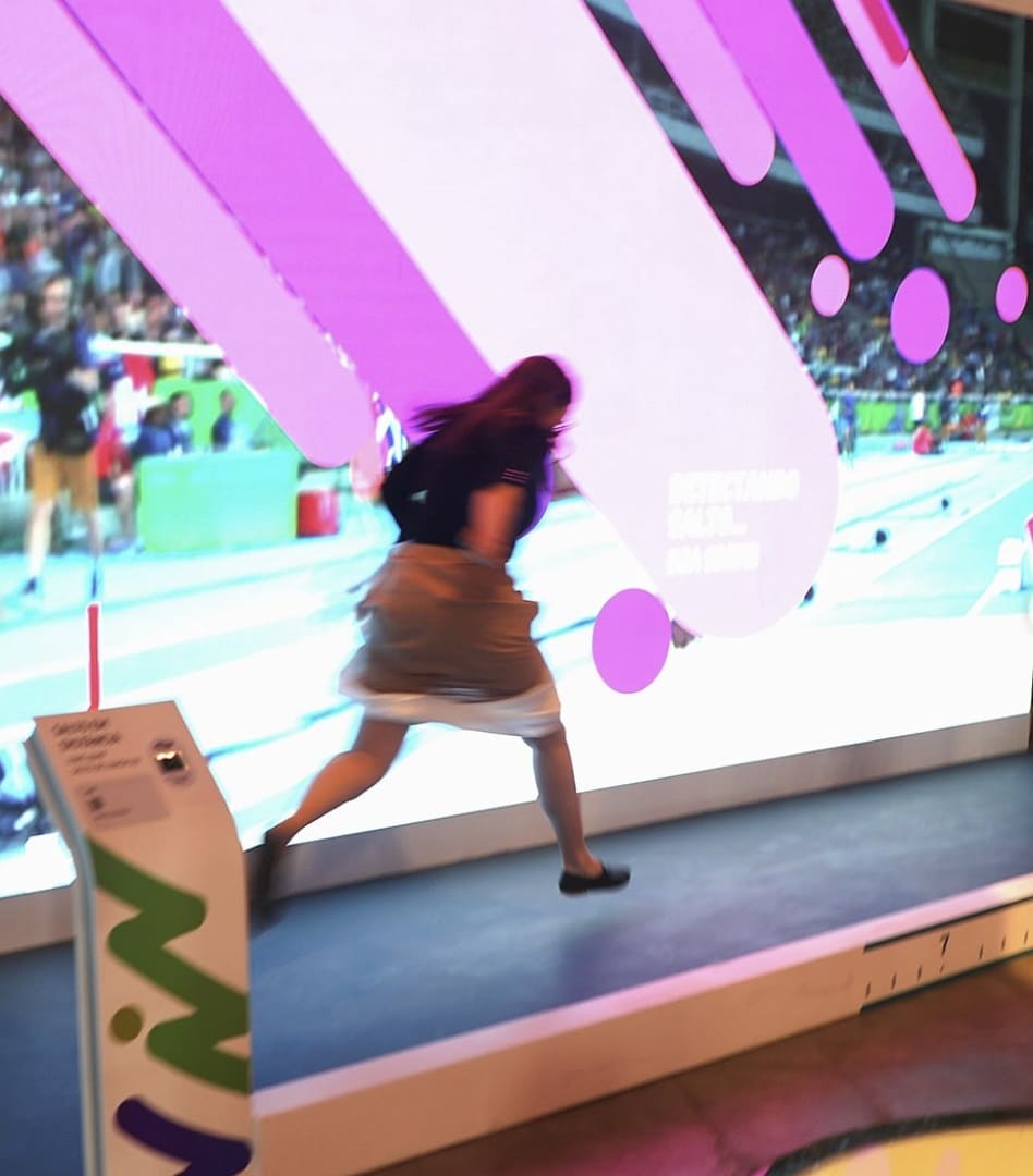 Uma pessoa é capturada em movimento rápido, saltando em frente a um grande painel digital interativo que exibe gráficos coloridos e uma cena de estádio de atletismo.