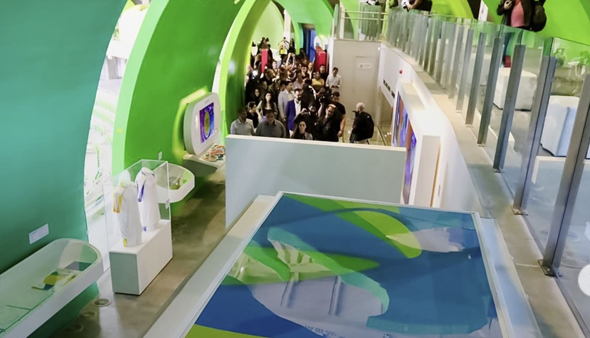 Vista aérea de um corredor moderno do Museu Olímpico com arcos verdes vibrantes, vitrines de uniformes e um grupo de pessoas ao fundo.
