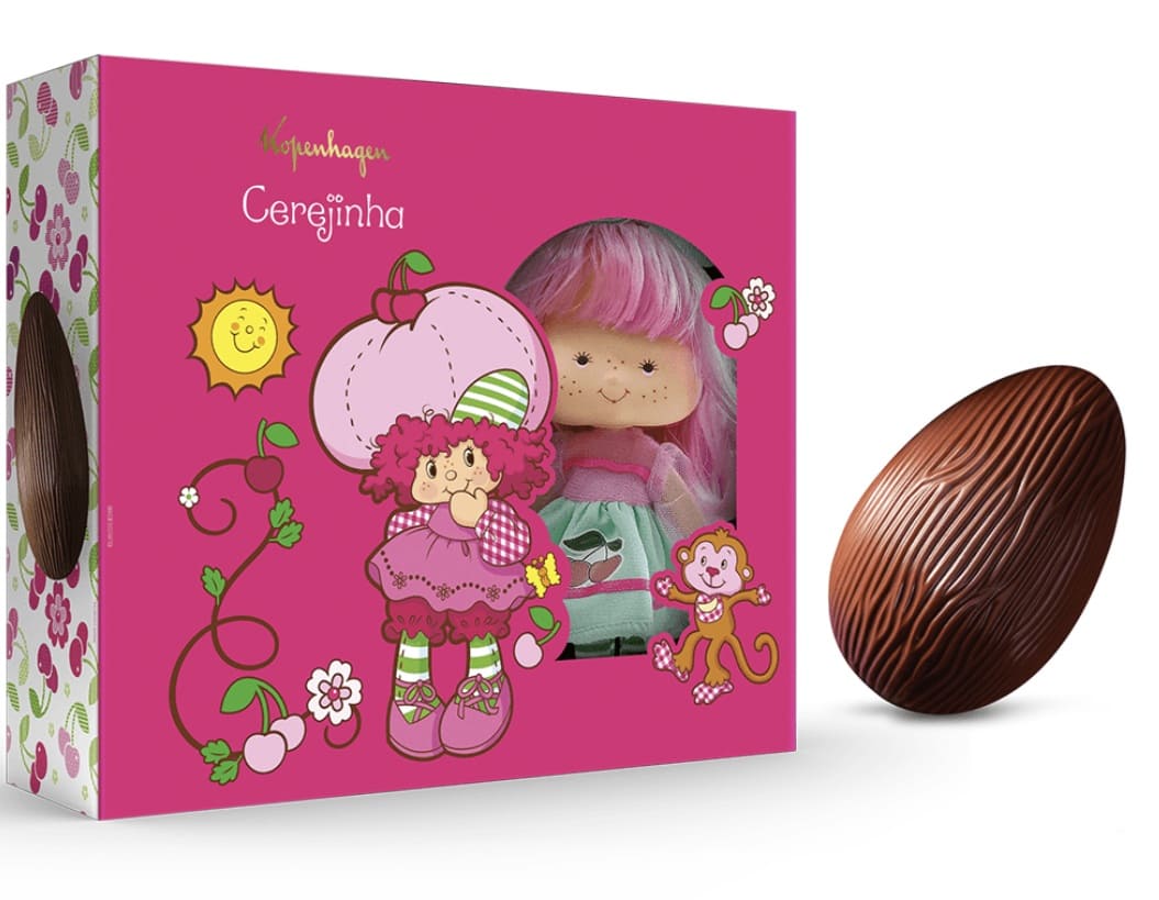 Embalagem rosa do Ovo de Páscoa Cerejinha da Kopenhagen mostrando a boneca colecionável em um visor transparente, acompanhada por um ovo de chocolate ao leite texturizado ao lado.