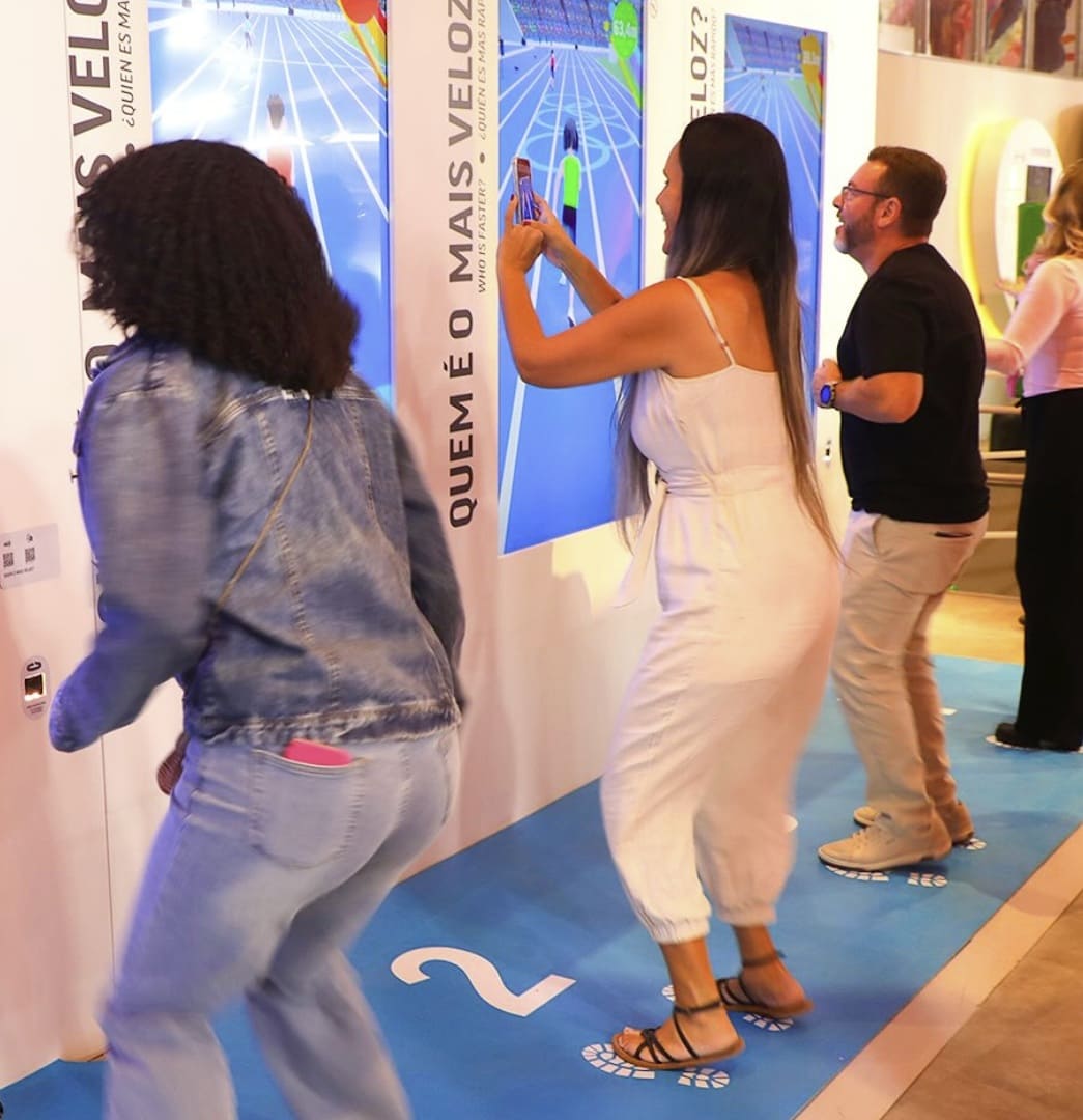 Três pessoas interagem de forma animada com uma tela digital interativa de atletismo no Museu Olímpico, em uma pista azul numerada.
