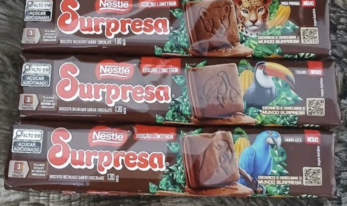 Foto em ângulo superior de três pacotes de biscoito Nestlé Surpresa (edição limitada) empilhados. As embalagens são marrons e destacam ilustrações de uma onça, um tucano e uma arara-azul, com o biscoito real posicionado à frente de cada desenho.