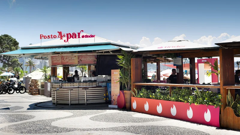 Fotografia de um quiosque na orla de Copacabana personalizado pela campanha do Tinder. O letreiro no topo diz "Posto 4 é par". O local possui detalhes em rosa e laranja, logos do Tinder e ombrelones brancos, situado sobre o calçadão de pedras portuguesas sob luz solar forte.