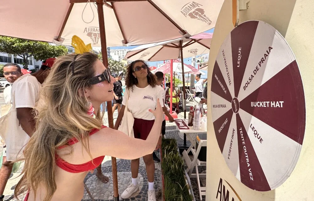 Mulher sorridente gira uma roleta de prêmios da Amarula em um evento de praia. A roleta exibe brindes como Bucket Hat, Bolsa de Praia e Leque.