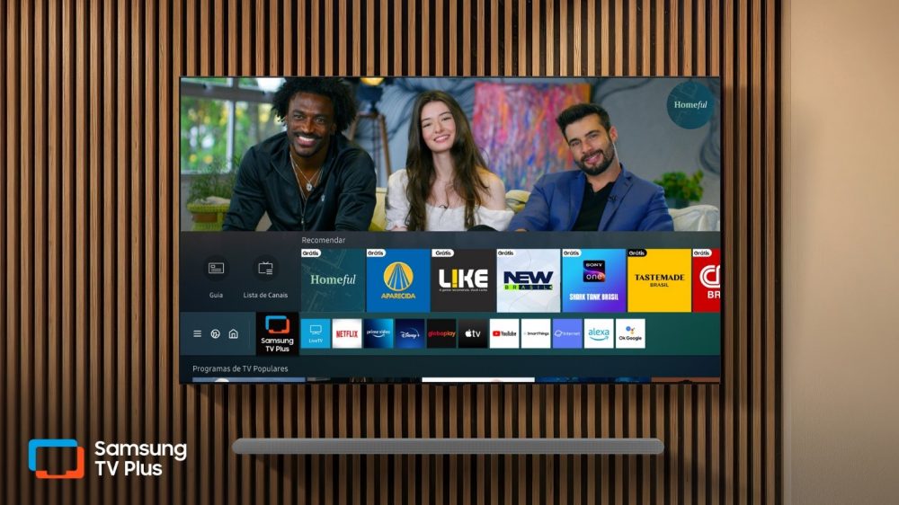 Uma Smart TV Samsung fixada em uma parede de ripas de madeira exibe a interface do Samsung TV Plus, mostrando opções de canais gratuitos e aplicativos de streaming.
