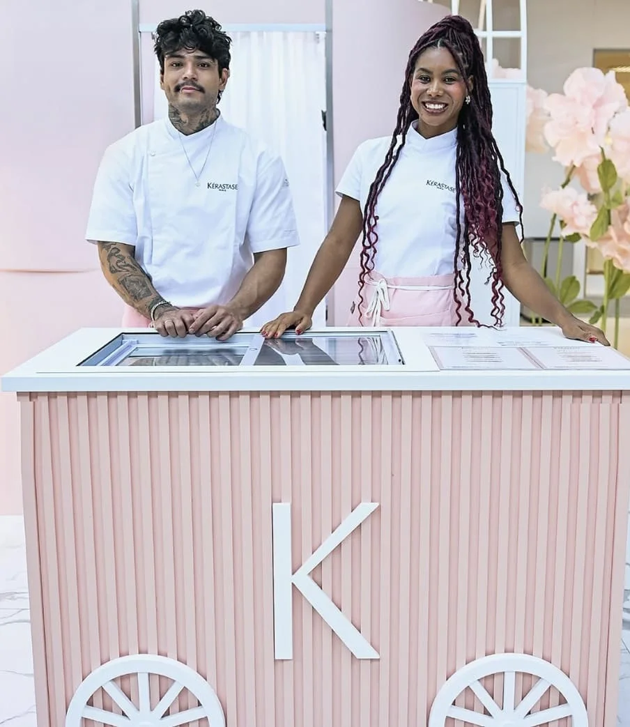 Duas pessoas sorridentes, vestindo uniformes brancos da Kérastase, posicionadas atrás de um carrinho de sorvete personalizado em tons de rosa com a letra "K" em destaque.
