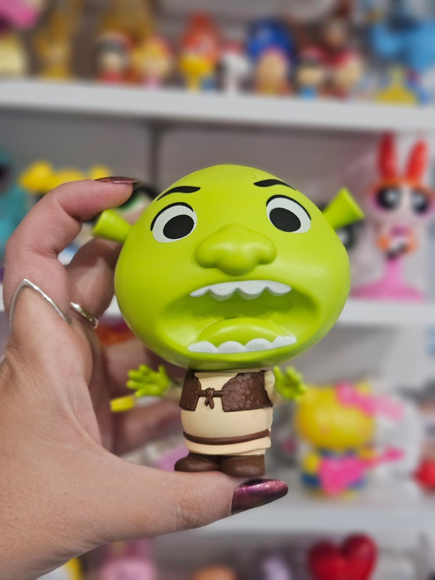 Uma mão segura um boneco colecionável do Shrek com cabeça grande e arredondada, olhos arregalados e boca aberta em uma expressão de surpresa. O boneco veste seu traje clássico de colete marrom e túnica bege.