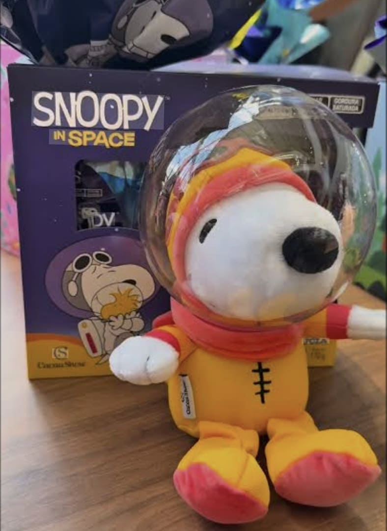 Close de uma pelúcia do Snoopy Astronauta fora da caixa, com traje espacial amarelo e laranja e capacete de acrílico transparente, posicionada à frente da embalagem temática da Cacau Show.
