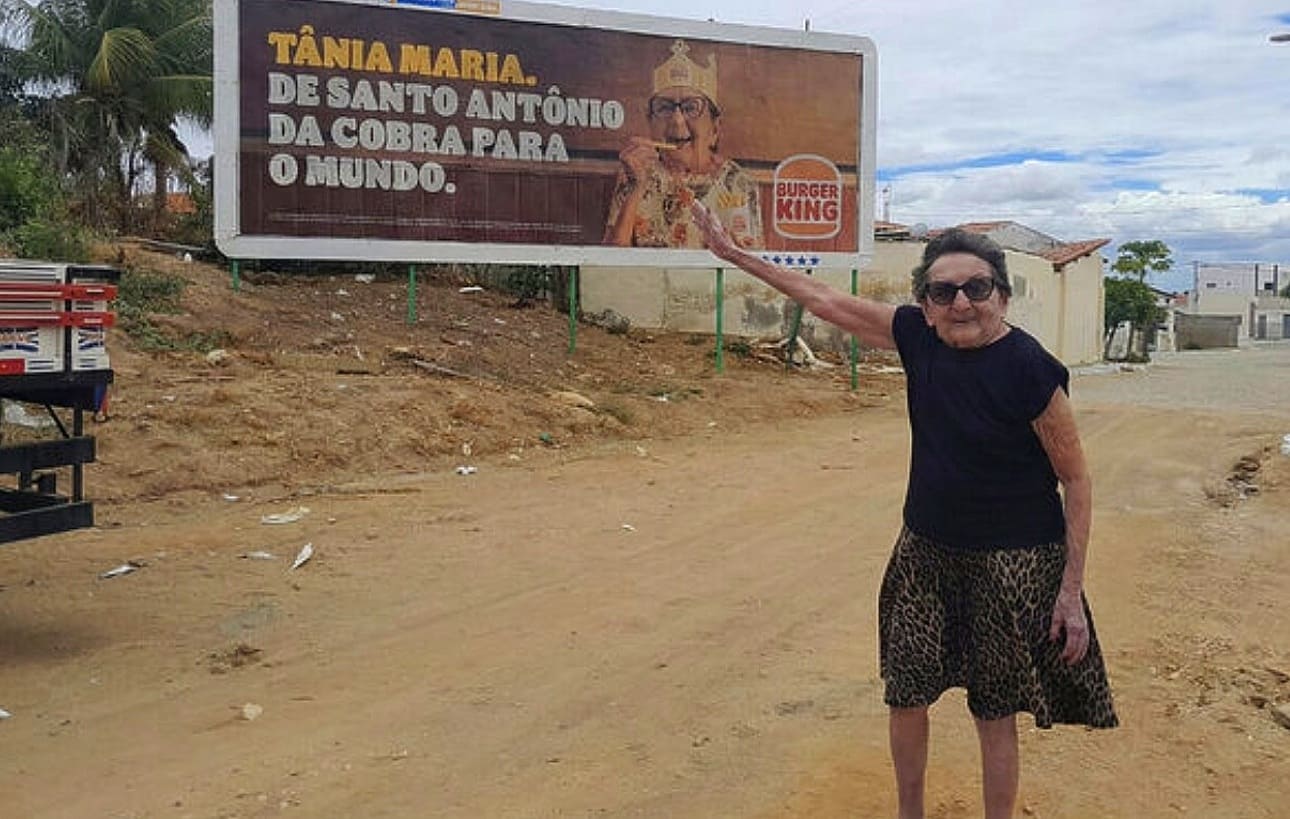 Uma senhora idosa, Tânia Maria, aponta com orgulho para um outdoor do Burger King que a homenageia. No outdoor, ela aparece sorridente com uma coroa da marca e o texto "De Santo Antônio da Cobra para o mundo". Ela está em uma rua de terra, usando óculos escuros e saia de estampa de oncinha.