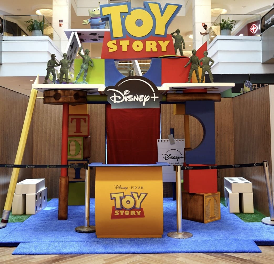 Foto frontal da entrada da experiência interativa Toy Story no Boulevard Shopping Belém. Inclui um balcão de atendimento amarelo, carpete azul e uma estrutura temática feita de blocos gigantes, dados e lápis.