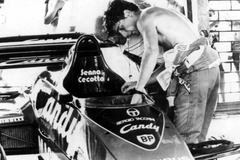 : Fotografia em preto e branco granulada, de close-up, de um jovem Ayrton Senna, sem camisa, debruçado sobre o cockpit de um carro de corrida Toleman na garagem dos boxes.