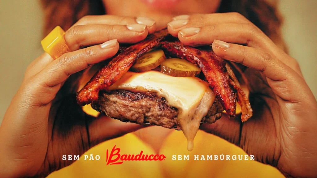 Uma pessoa segura um disco de carne de hambúrguer grelhado com queijo derretido, picles e bacon por cima, sem o pão de hambúrguer. Texto abaixo: "Sem pão Bauducco sem hambúrguer".
