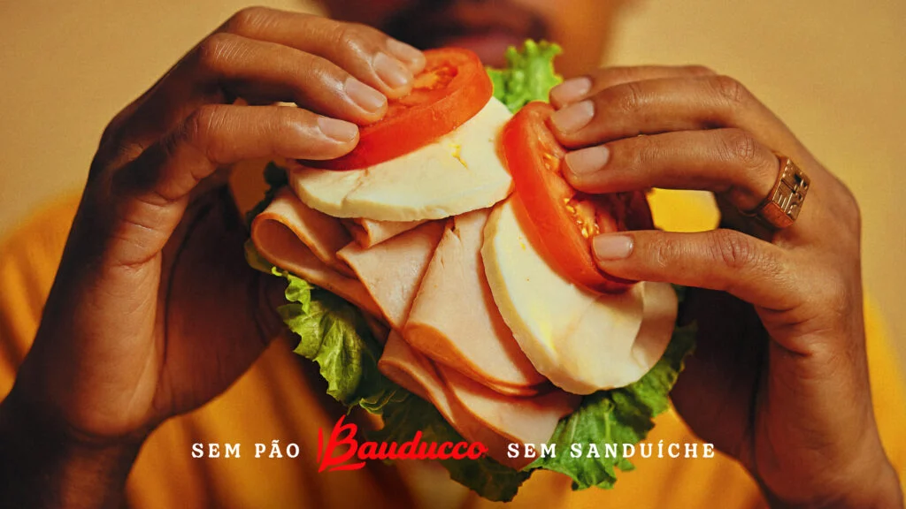 Mãos segurando uma pilha de fatias de peito de peru, queijo, tomate e alface. Não há pão envolvendo os ingredientes. Texto abaixo: "Sem pão Bauducco sem sanduíche".