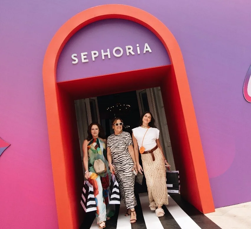 Uma foto de três mulheres sorrindo enquanto caminham para fora de um grande portal arqueado de cor roxa e coral. O letreiro "SEPHORiA" está acima do arco. Elas seguram sacolas de compras da Sephora.