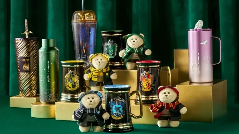 Exposição da coleção Harry Potter x Starbucks sobre pedestais dourados, apresentando garrafas térmicas, copos com canudo, canecas pretas com brasões das casas e ursos Bearista vestidos com uniformes de Hogwarts.