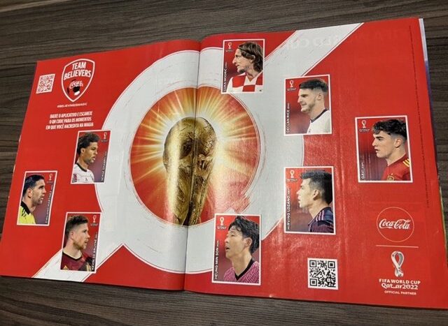 Página de álbum de figurinhas aberta sobre uma mesa de madeira. O fundo é vermelho com um grande círculo central branco irradiando luz dourada ao redor de uma imagem da taça da Copa do Mundo FIFA. O topo esquerdo tem o escudo "Team Believers Coca-Cola" e um QR code. Sete figurinhas de jogadores, incluindo Luka Modrić e Son Heung-min, estão coladas na página. O logo da Copa do Mundo Catar 2022 está no canto inferior direito.
