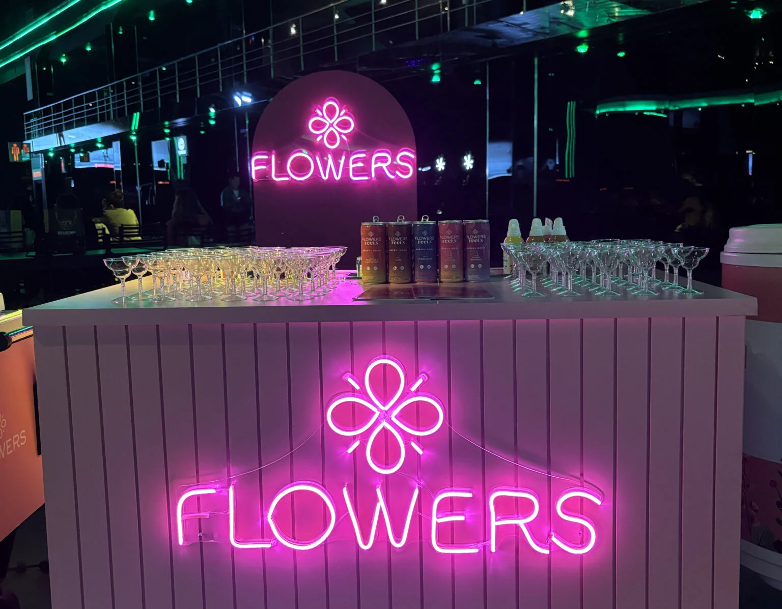Balcão de eventos iluminado com letreiros em neon rosa da marca "Flowers", com taças de cristal e latas de bebida organizadas sobre o tampo branco.