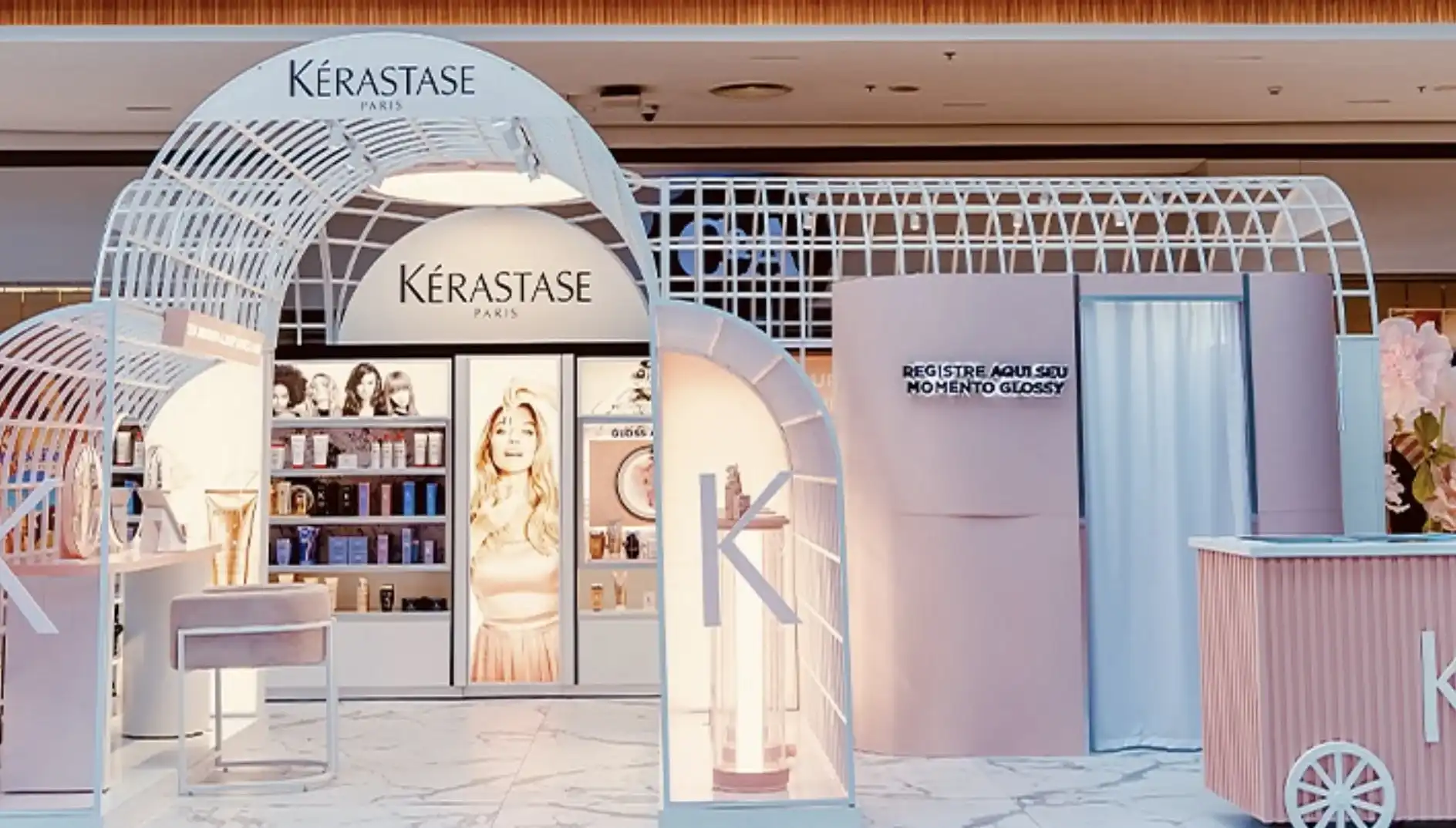 Foto de um estande da Kérastase em um shopping com arquitetura inspirada em uma estufa metálica branca e arredondada. O espaço possui tons de rosa suave e branco, incluindo uma bancada de atendimento, prateleiras com produtos, uma poltrona de veludo e uma cabine com a frase 'Registre aqui seu momento Glossy'.