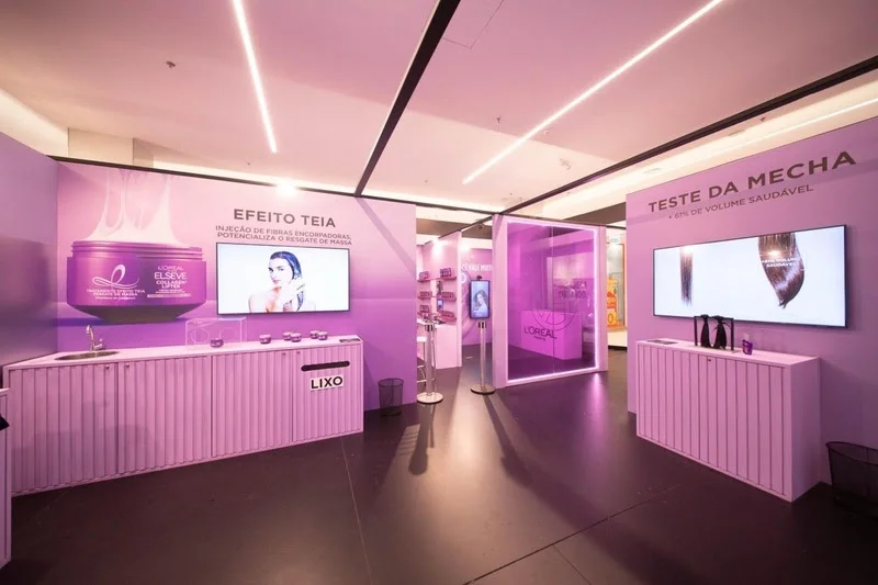Interior de um estande da L'Oréal Paris decorado em tons de lilás, com estações de teste de produtos. Painéis destacam o 'Efeito Teia' e o 'Teste da Mecha' da linha Elseve Collagen Lifter, com telas exibindo vídeos demonstrativos.