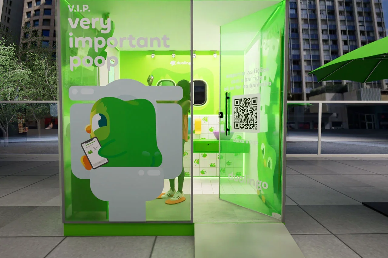 Vista frontal de uma cabine de banheiro móvel e transparente em tons de verde vibrante, instalada em uma área urbana ao ar livre. Na fachada de vidro, há a ilustração da coruja Duo sentada em um vaso sanitário mexendo no celular, acompanhada do texto "V.I.P. Very Important Poop". Uma porta de vidro semiaberta revela um código QR e o interior decorado.