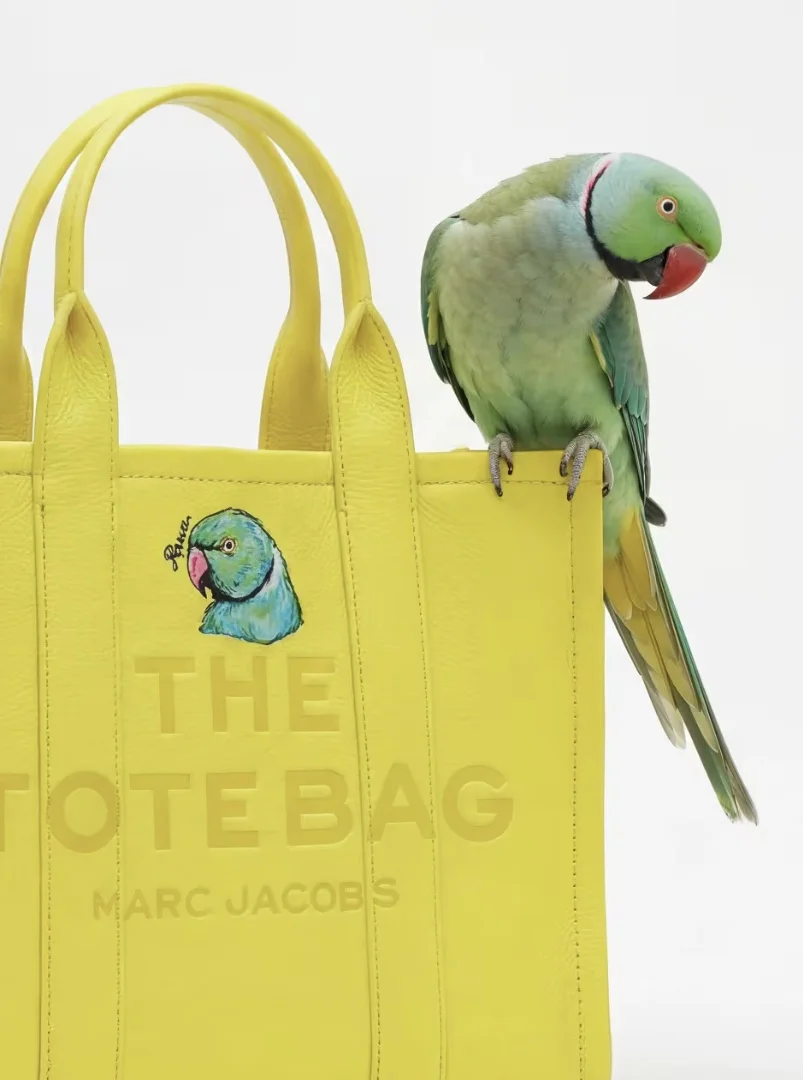 Uma sacola de compras amarela vibrante "The Tote Bag Marc Jacobs" com uma ilustração de periquito. Um periquito-de-colar vivo está empoleirado na borda.