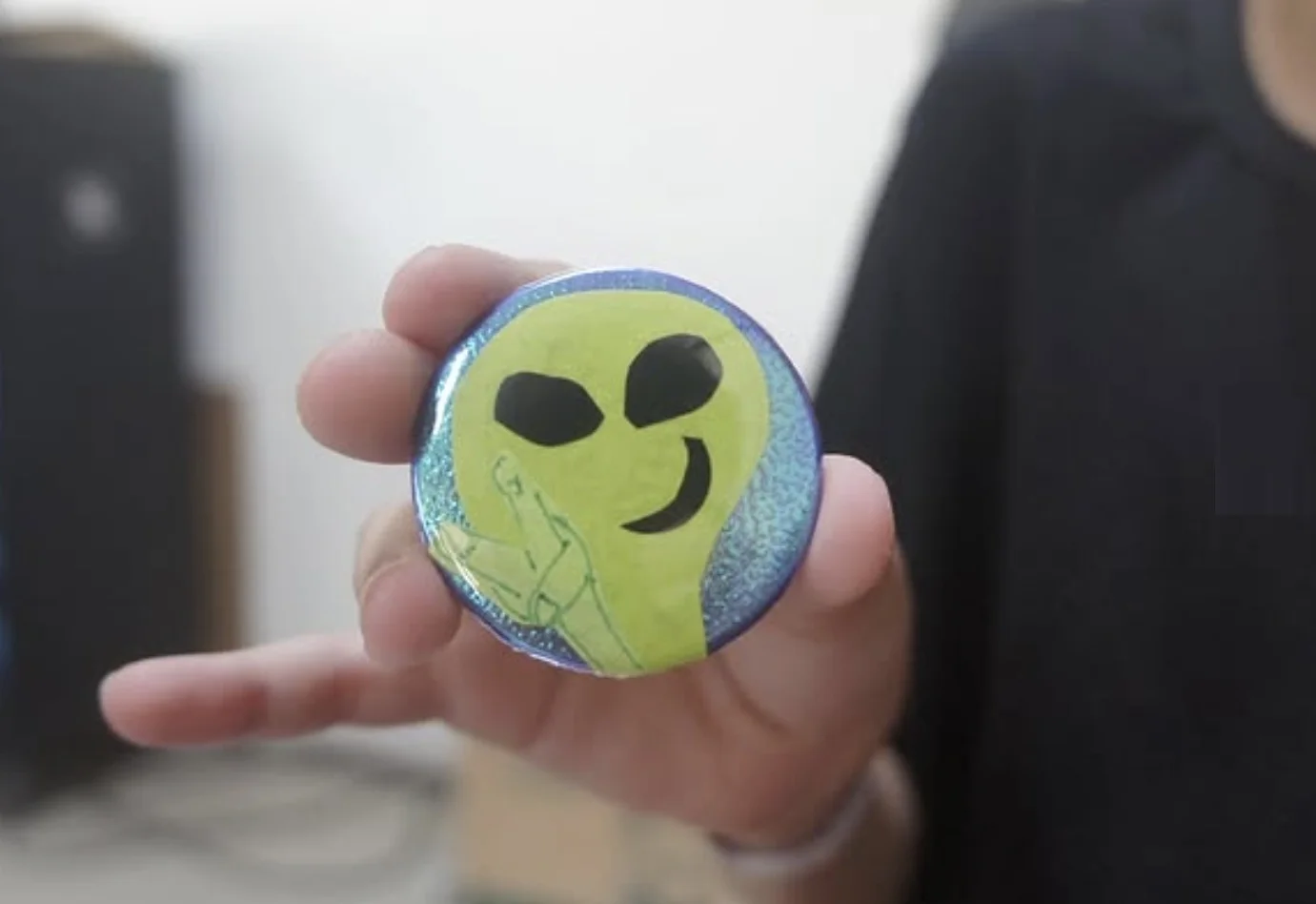 Uma mão segurando um bóton redondo com a ilustração de um alienígena verde sorridente fazendo um sinal de paz, sobre um fundo azul brilhante.