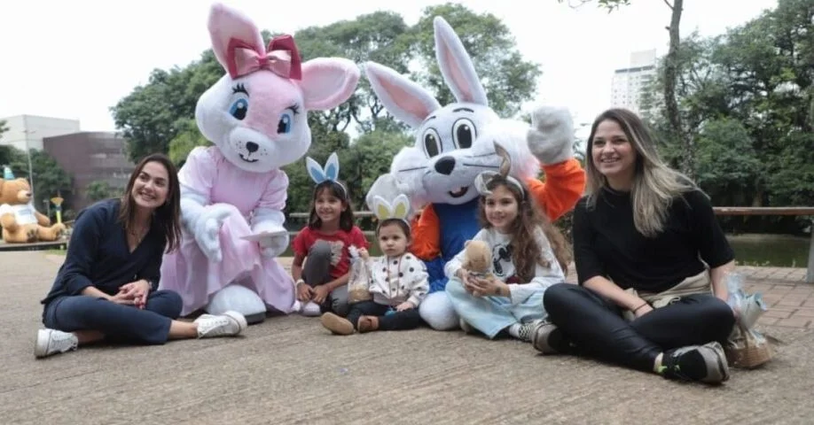 Duas mulheres e três crianças posam sentadas no chão ao lado de dois personagens de coelho da Páscoa em um ambiente arborizado.