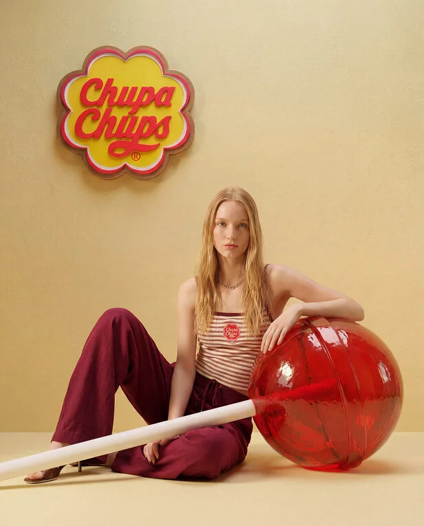 Modelo sentada no chão ao lado de um pirulito vermelho gigante em um cenário bege com o logotipo da Chupa Chups na parede.