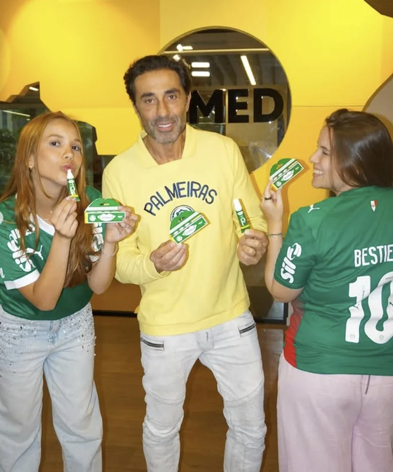 Três pessoas posando com os produtos da linha Carmed Palmeiras em um ambiente com o logo da Cimed ao fundo.