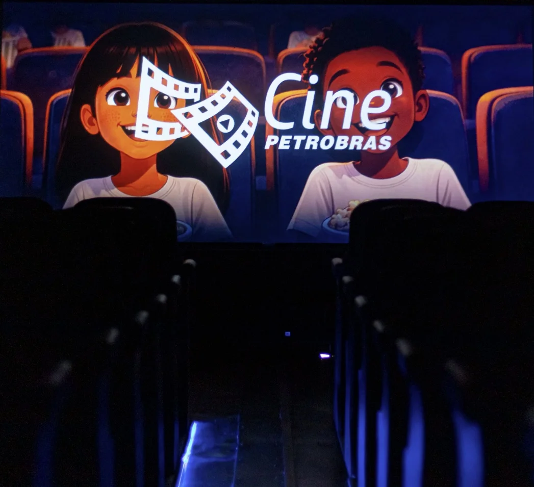 Interior de uma sala de cinema escura com poltronas pretas em silhueta. Na tela iluminada, uma ilustração exibe duas crianças sorridentes segurando pipoca ao lado do logotipo branco "Cine Petrobras".