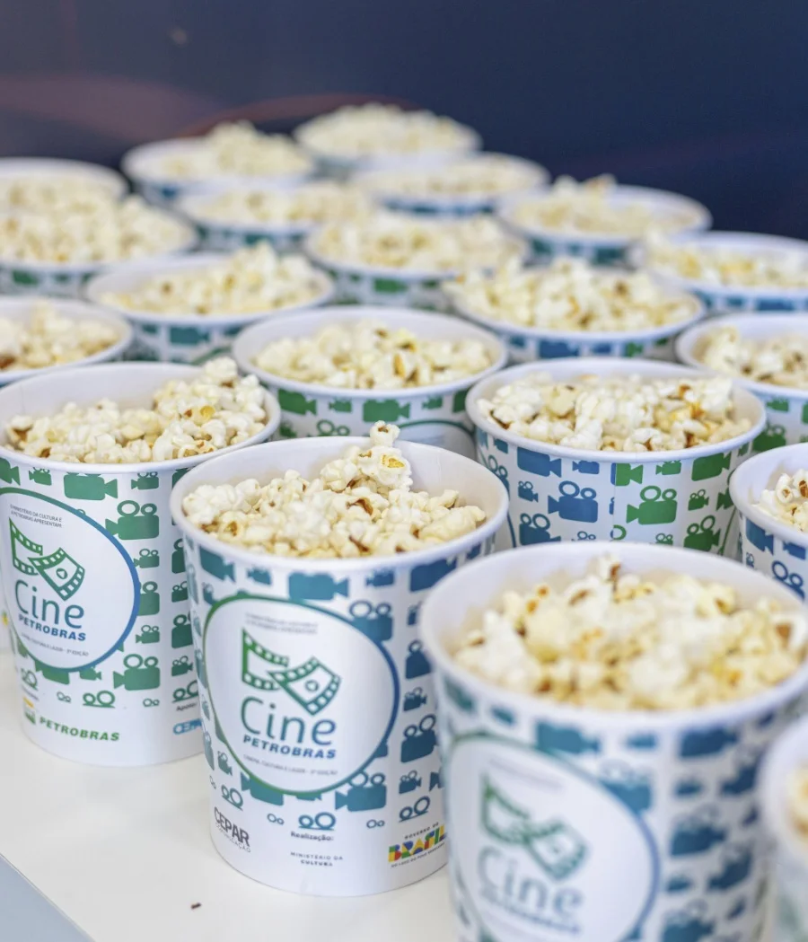 Close em vários baldes brancos cheios de pipoca, organizados em fileiras. Os baldes possuem a logomarca do "Cine Petrobras" e ícones de câmeras de cinema em tons de verde e azul.