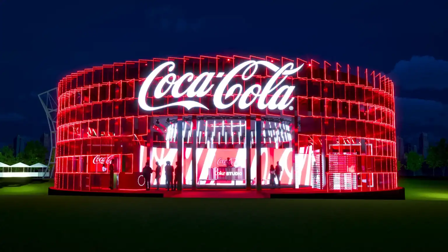 Estrutura circular iluminada em vermelho vibrante da ativação "Coke Studio" da Coca-Cola em um festival noturno, com o logotipo da marca em destaque no topo e pessoas circulando em seu interior.