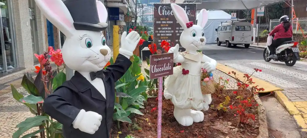Duas grandes estátuas de coelhos de Páscoa, vestindo roupas de gala e noiva, ao lado de uma placa anunciando um "Festival de Chocolate" na rua.Duas grandes estátuas de coelhos de Páscoa, vestindo roupas de gala e noiva, ao lado de uma placa anunciando um "Festival de Chocolate" na rua.