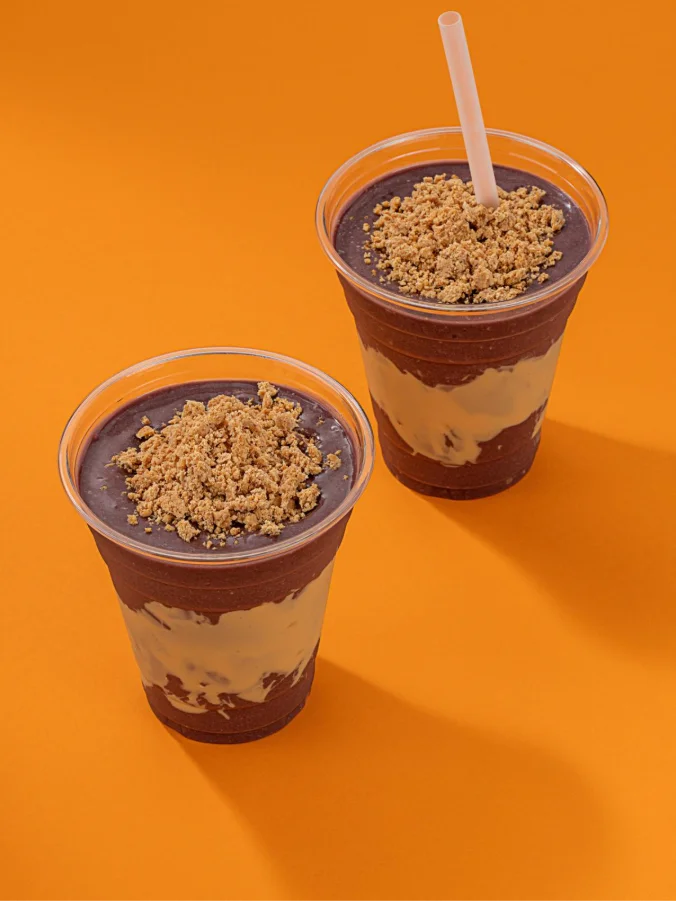 Dois copos plásticos de açaí com uma camada generosa de creme de amendoim ou paçoca cremosa no meio e farofa de paçoca por cima, sobre um fundo laranja vibrante.