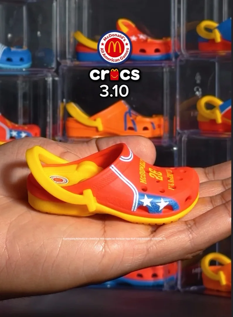Close-up de uma mão segurando um mini Crocs colecionável nas cores vermelho e amarelo, com detalhes de estrelas e o número 26. Ao fundo, outros modelos similares aparecem organizados em caixas acrílicas transparentes sob o logotipo da parceria McDonald’s e Crocs.