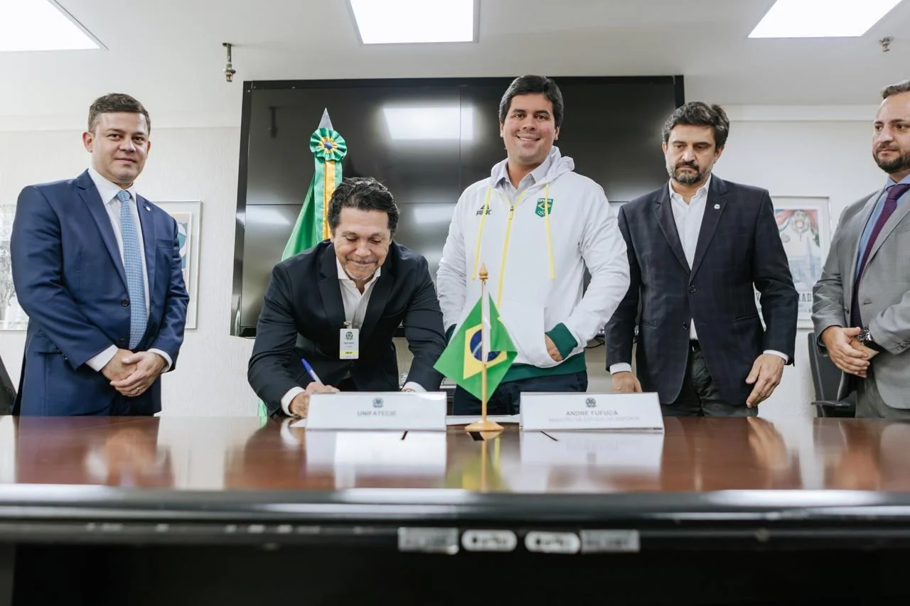 Autoridades brasileiras reunidas em uma mesa oficial durante a assinatura de um documento. Ao centro, um homem assina papéis enquanto o Ministro do Esporte, André Fufuca, observa ao lado de outros representantes.