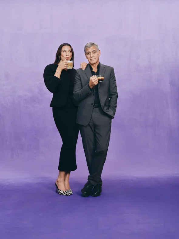 Retrato de estúdio de Dua Lipa e George Clooney posando juntos contra um fundo roxo. Ela está à esquerda com um vestido preto, segurando uma caneca de latte, e ele à direita de terno cinza, segurando uma xícara de café preto.