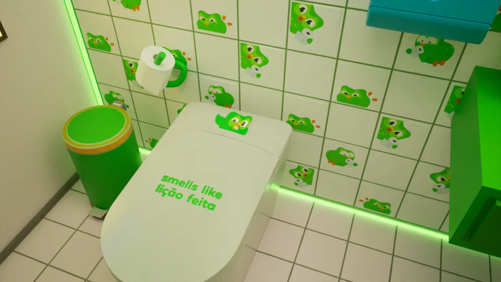 Close de um vaso sanitário branco em um banheiro temático do Duolingo. Na tampa, lê-se a frase "smells like lição feita" em letras verdes. Ao redor, azulejos brancos com adesivos da coruja Duo, uma lixeira verde e iluminação em LED neon verde no rodapé.