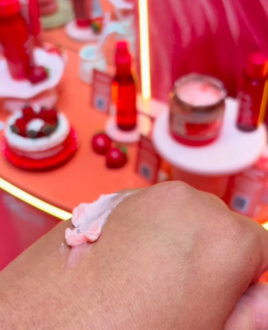 Close-up da mão de uma pessoa testando um creme rosado, com diversos produtos de beleza vermelhos e uma decoração temática de morango ao fundo.