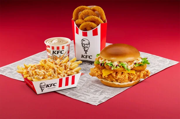 Combo do KFC sobre fundo vermelho, composto por um sanduíche de frango crocante com anéis de cebola, batatas fritas com cobertura, uma porção extra de onion rings e um molho.