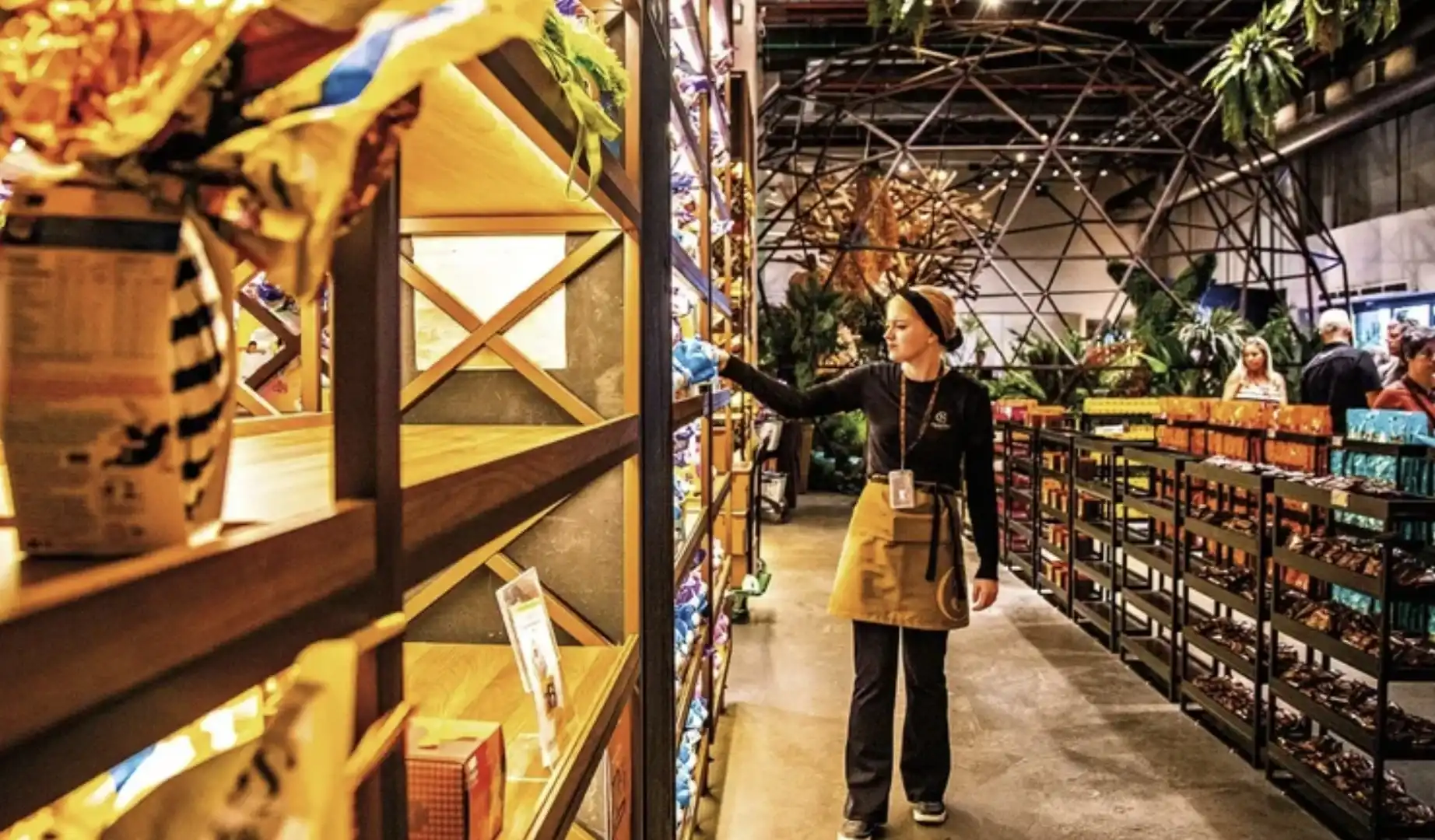 Funcionária organizando prateleiras em uma loja da Cacau Show com design moderno e elementos de natureza.