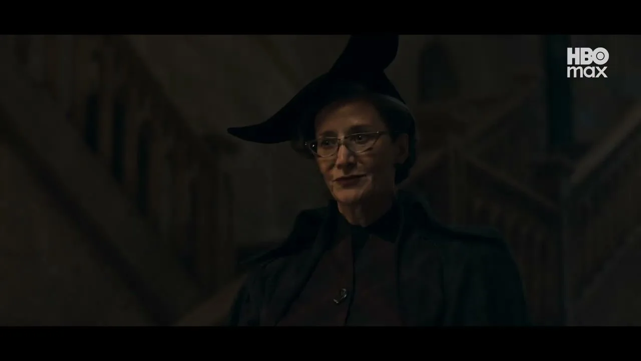 Foto em plano médio de uma mulher de meia-idade com cabelos grisalhos presos sob um chapéu de bruxa pontudo e torto, usando óculos de armação fina e um casaco escuro. Ela tem uma expressão severa. O fundo é um grande salão gótico ou escadaria, com o logotipo da HBO Max no canto superior direito.