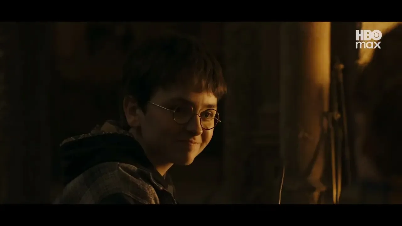 Close em close-up de perfil de um jovem ator com óculos redondos e uma leve cicatriz de raio na testa, olhando para a direita e sorrindo. Ele usa uma jaqueta xadrez escura. O fundo é mal iluminado e embaçado, com o logotipo da HBO Max no canto superior direito.