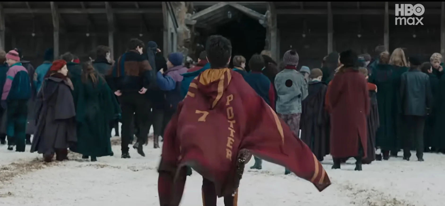 Uma cena de um filme, vista por trás, mostra o personagem principal, de costas, correndo em direção a uma multidão de pessoas reunidas na neve diante de um grande celeiro de madeira. Ele veste uma capa de Quadribol do Grifinória, marrom-avermelhada, com o número "7" e o nome "POTTER" em letras douradas. O logotipo "HBO Max" está no canto superior direito.